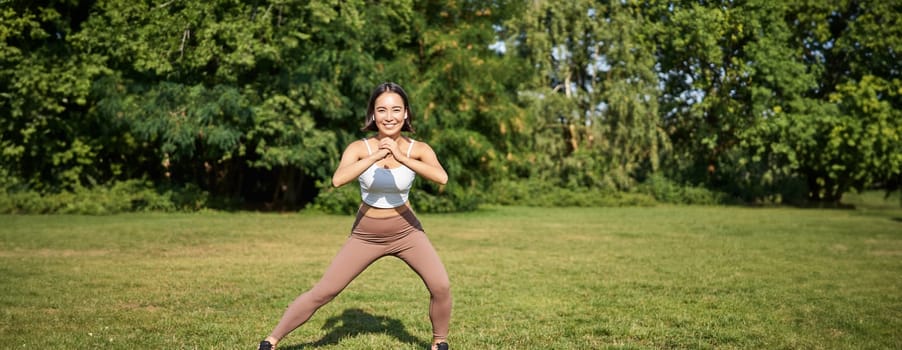 Wellbeing and sport complex. Young asian woman stretching, doing squats and workout on fresh air, smiling pleased.