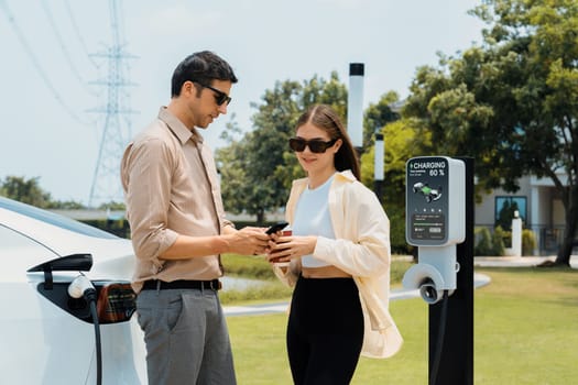 Young couple with coffee pay for electricity while recharge EV car battery at charging station connected to electrical power grid tower as electric industrial for eco friendly car travel. Expedient