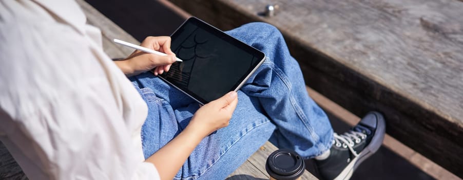 Cropped shot of female hands drawing on digital graphic tablet with pen, sitting with cup of coffee outdoors on bench.