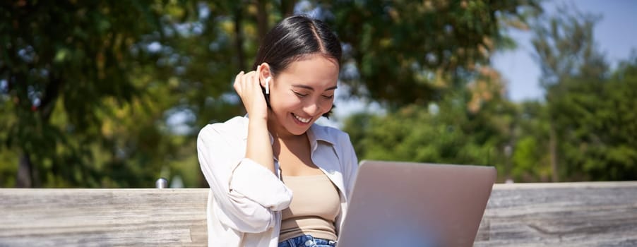 Beautiful asian girl laughing, watching video on laptop, listening music or video chatting online while sitting in park on bench.