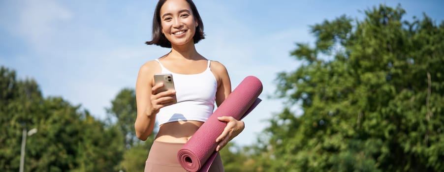 Portrait of young asian woman checking her phone during workout, walking in park with rubber mat, going to the gym, holding smartphone.