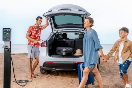 Family vacation trip traveling by the beach with electric car, lovely family taking luggage out while charging EV car battery with clean energy. Alternative family travel by eco-friendly car.Perpetual