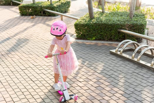 Child riding scooter. Happy Asian little kid girl wear safe helmet playing pink kick board on road in park outdoors on summer day, Active children games outside, Kids sport healthy lifestyle concept