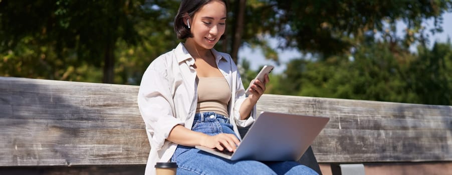 Young asian girl talking on mobile phone, using laptop, working online from park, sitting on bench on sunny day with computer.