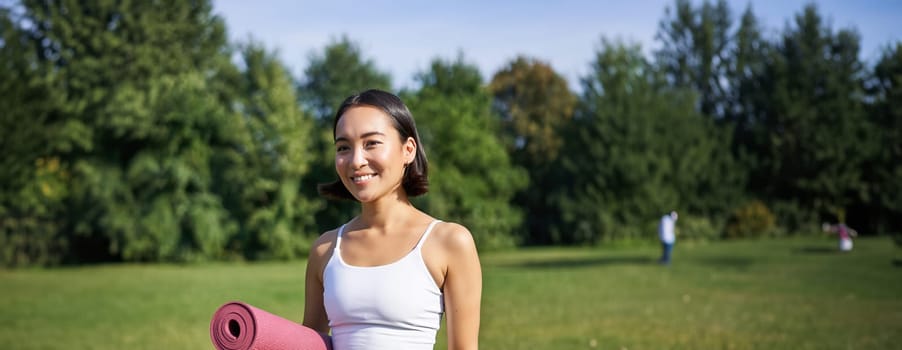 Vertical portrait of young asian fitness girl walks with rubber mat for yoga, goes on training session on fresh air in park, wears sports clothing.