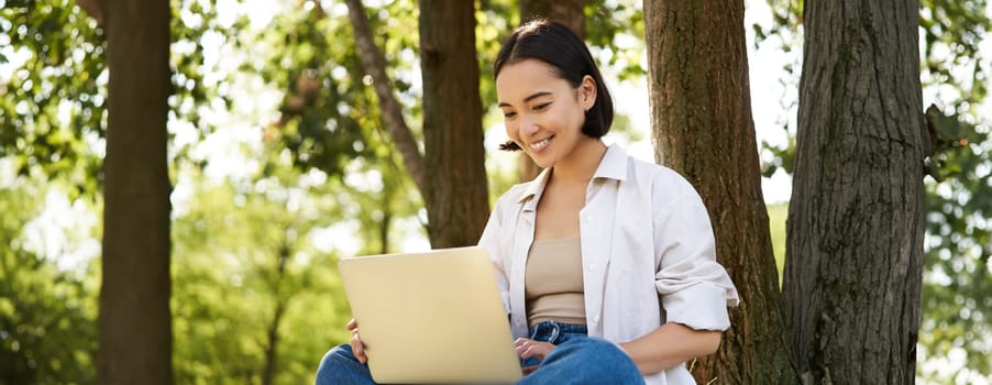 Happy young asian girl sits in park near tree, looking at laptop, working remotely from outdoors, talking to someone, video chat.