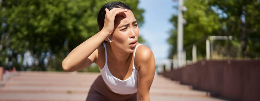 Tired young female runner, asian girl taking break during workout, stop jogging, panting while breathing, running in park.