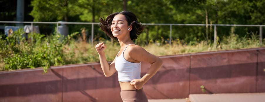 Wellbeing. Smiling asian fitness girl, runner in park, smiling and running, doing jogging workout outdoors on streets.