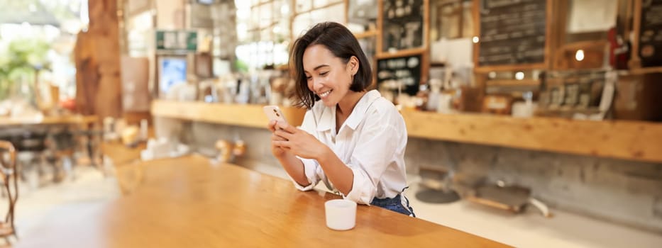 Vertical portrait of stylish asian woman sitting in cafe, drinking coffee and using smartphone. Lifestyle and people concept