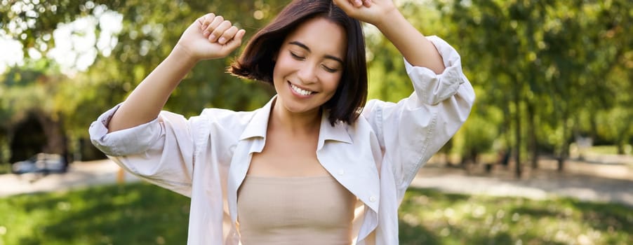 People lifestyle. Portrait of young brunette woman dancing, smiling and laughing, walking in park with hands lift up high, enjoying summer day outside.