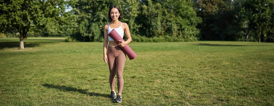 Portrait of fitness girl walking in park with rubber yoga mat, going on sports training outdoors on lawn.