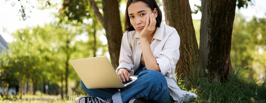 Portrait of young asian woman sitting in park near tree, working on laptop, using computer outdoors.