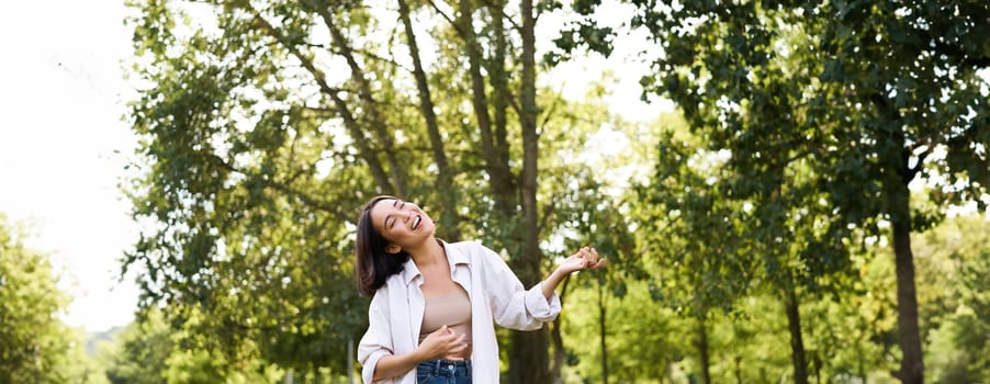 Happy young asian woman walking alone, dancing and singing in park, smiling carefree. People and lifestyle concept.