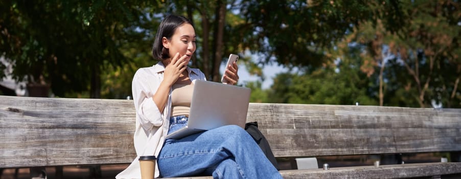 Asian girl looks surprised at smartphone screen, checking mobile phone notifications and looking excited, sitting with laptop in park on bench.