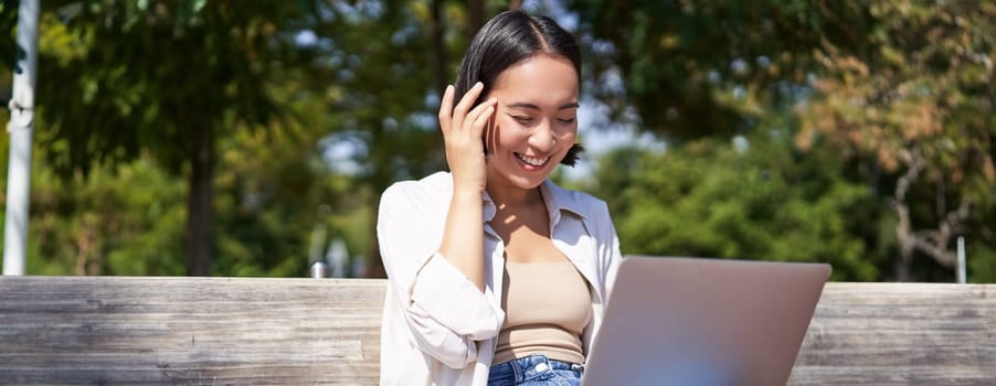 Beautiful asian girl laughing, watching video on laptop, listening music or video chatting online while sitting in park on bench.