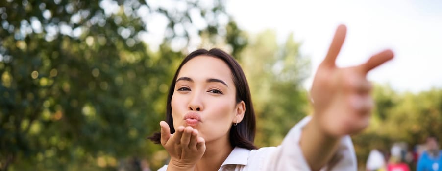 Smiling asian girl takes selfie, video chats, holds smartphone and speaks at camera, posing in park.