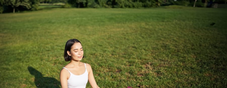 Mindfulness and meditation. Young asian woman smiling while doing yoga, relaxing in asana on rubber mat, doing exercises in park on fresh air.