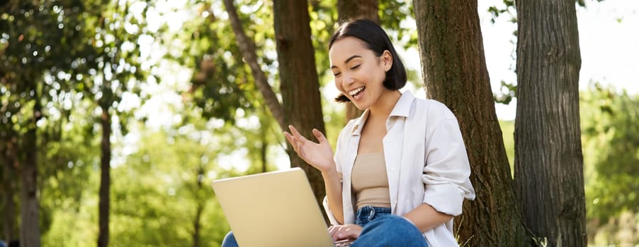 Portrait of asian woman sitting on grass near tree, using laptop, working, doing homework remotely on sunny summer day.