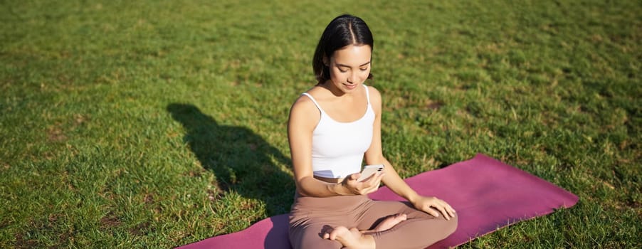 Asian girl follows yoga training app, looking at smartphone, meditating and doing exercises on fresh air in park.