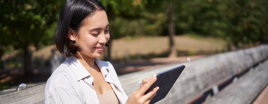 Portrait of young asian woman drawing on fresh air in park, sitting with graphic tablet and digital pen, smiling happy.