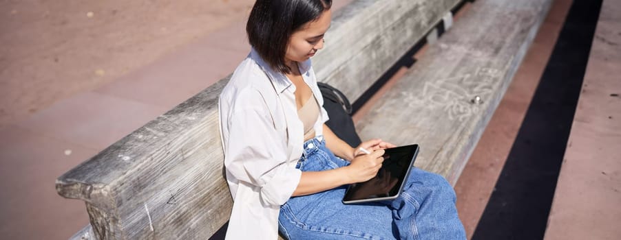 Portrait of young artist, asian girl drawing in digital tablet with graphic pen, sitting outdoors in park, getting inspiration.