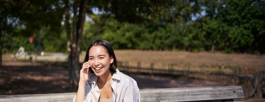 Portrait of happy young asian woman talking on mobile phone in park, sitting on bench and having a telephone call, chatting lively.