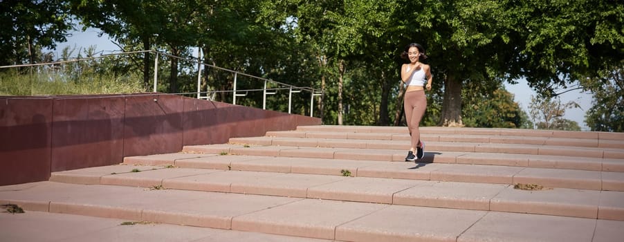 Healthy fitness girl running outdoors on street, wearing uniform, jogging on fresh air and listening music in wireless headphones.