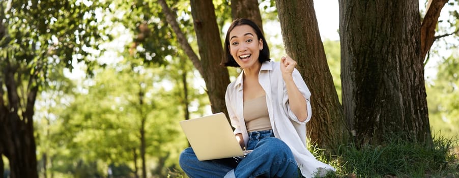 Enthusiastic young asian girl, sitting with laptop beside tree in green sunny park, celebrating, triumphing and smiling.
