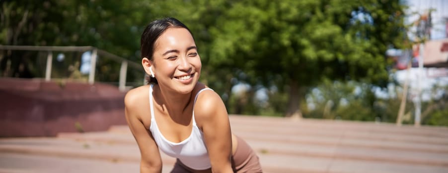 Portrait of asian woman taking break, breathing heavily and panting after running, jogger standing and wiping sweat off forehead, smiling pleased.