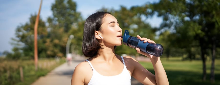 Happy asian sportswoman, runner drinks water from bottle while running, workout on fresh air in park.