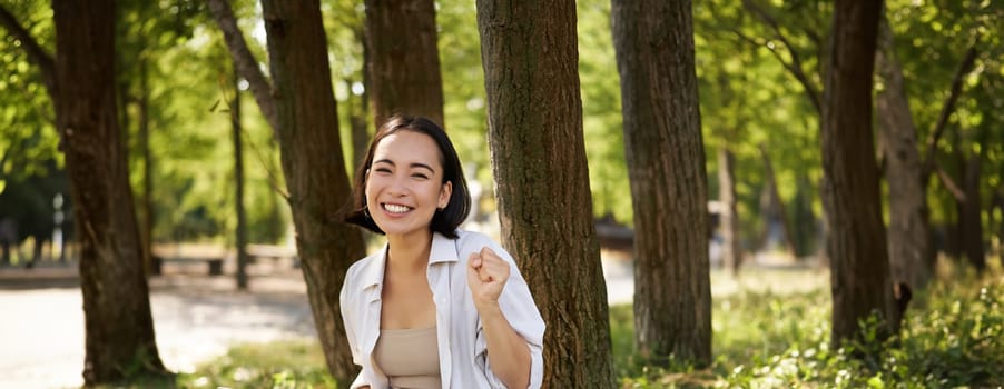 Enthusiastic young asian girl, sitting with laptop beside tree in green sunny park, celebrating, triumphing and smiling.