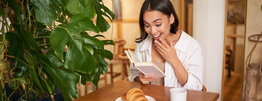 Asian girl looks amazed at pages, reading with excitement, sitting in cafe and eating croissant.