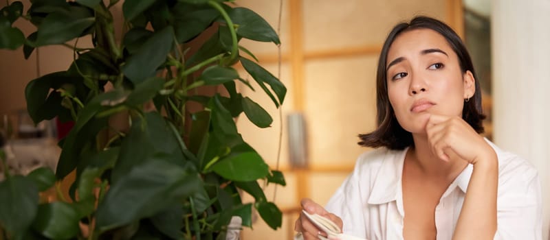 Vertical shot of asian girl sits alone in cafe, reads book and looks upset, drinks coffee with croissant.