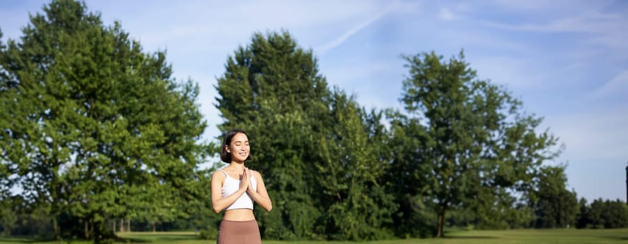 Young mindful woman, meditating on fresh air, doing yoga exercises in park, standing on rubber mat in sportswear.