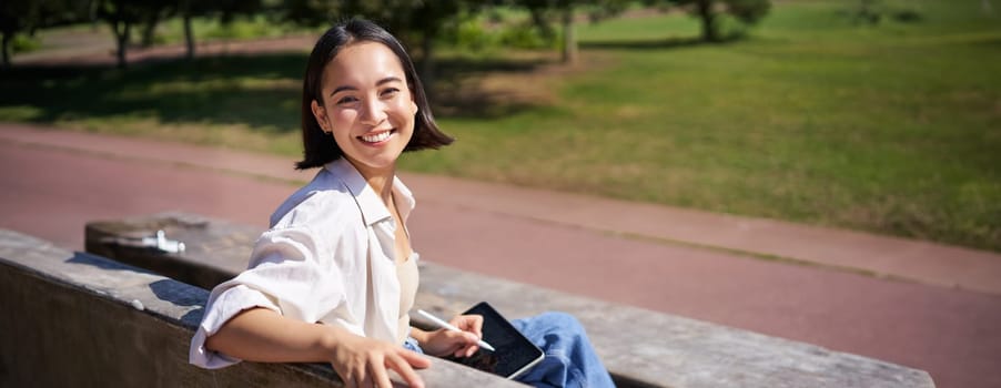 Happy korean woman sitting on bench with digital tablet and graphic pen, smiling, turning back at camera and talking lively.