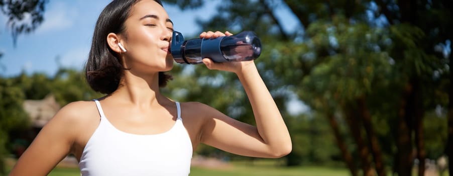Fit asian sportswoman drinks water while running marathon. Fitness girl jogging in park, staying hydrated. Copy space