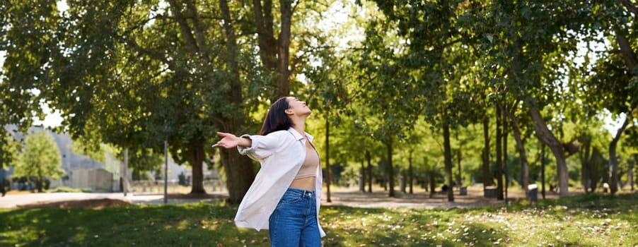 Carefree asian girl dancing, feeling happiness and joy, enjoying the sun on summer day, walking in park with green trees.