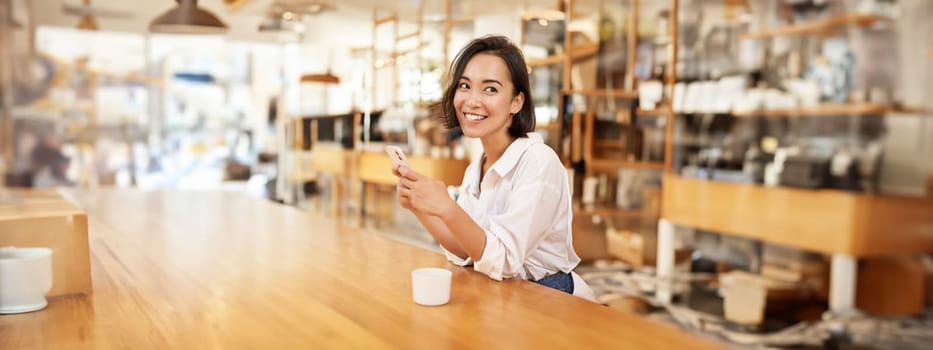 Vertical portrait of stylish asian woman sitting in cafe, drinking coffee and using smartphone. Lifestyle and people concept