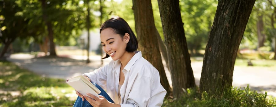 Beautiful young asian girl, student sits in park under tree and reading book, smiling, enjoying warm summer day outdoors.