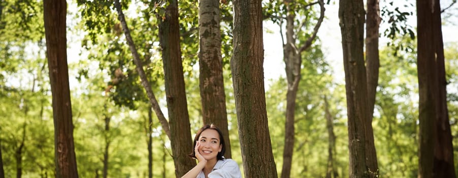 Woman sitting in park with her favourite book, leaning on tree under shade on sunny day, enjoying nature and calm relaxing atmosphere.