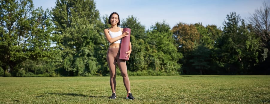 Confident and sporty young asian woman, standing in park with rubber mat, wearing sportswoman, smiling at camera, workout on fresh air.