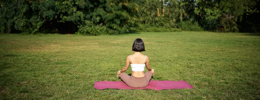 Rear view of woman silhouette doing yoga, sitting on fitness mat and meditating on green lawn.