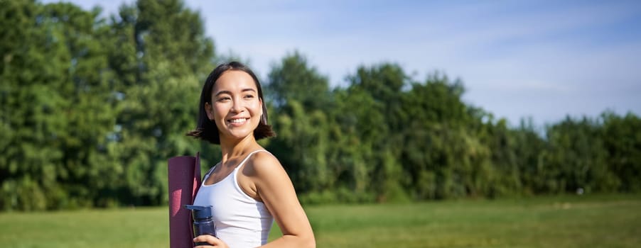 Vertical shot of fit and healthy asian woman posing in park, holding water bottle and yoga rubber mat for workout outdoors.