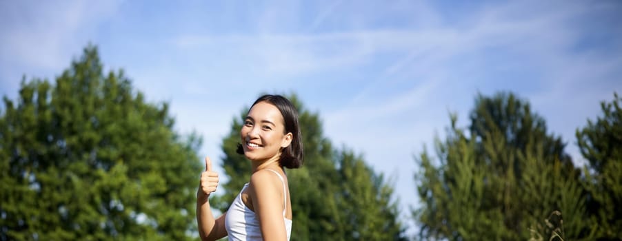 Portrait of asian woman say yes to yoga training in park, makes thumb up, sitting and meditating.