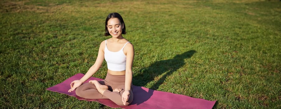 Portrait of young mindful woman, practice yoga, exercising, inhale and exhale on fresh air in park, sitting on rubber mat.