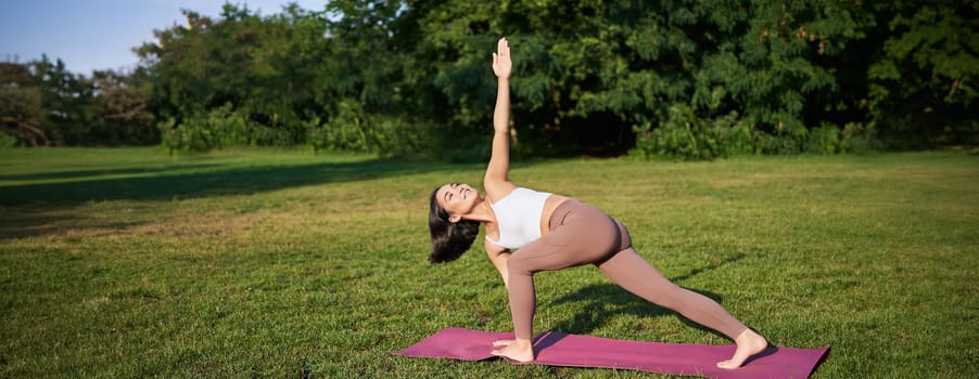 Vertical shot of young korean woman doing yoga training on rubber mat, making asana exercises on fresh air in park.