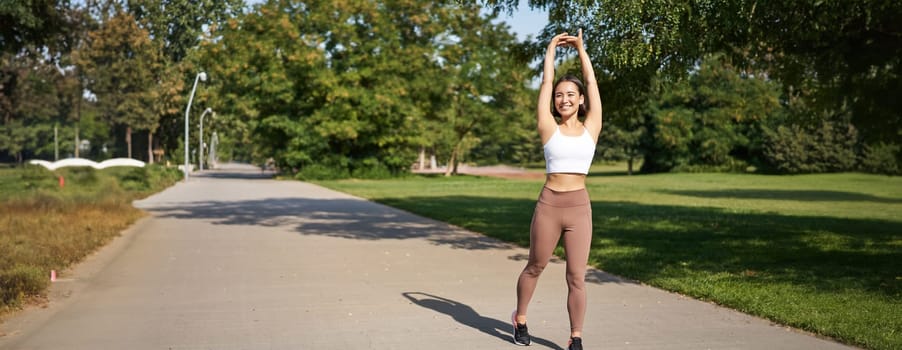 Smiling Asian girl stretching after good workout in park, listening music in wireless headphones, jogging outdoors.
