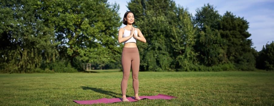 Young asian woman in sportswear, holding hands in namaste gesture, practice yoga, meditating and relaxing on fresh air in park, concept of mindfulness and wellbeing.