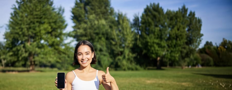 Vertical shot of asian woman shows thumbs up, recommending yoga training online, meditation app, doing exercises on fresh air in park, showing smartphone screen.