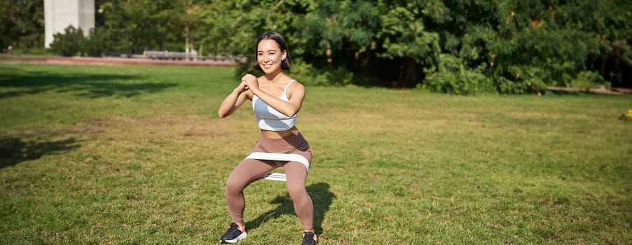 Woman using stretching band, resistance fitness rope for workout in park, doing squats, training on fresh air.